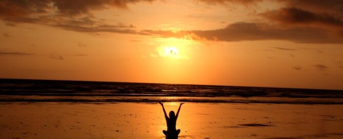 lady sitting on the beach with her hands ups watching the sunset